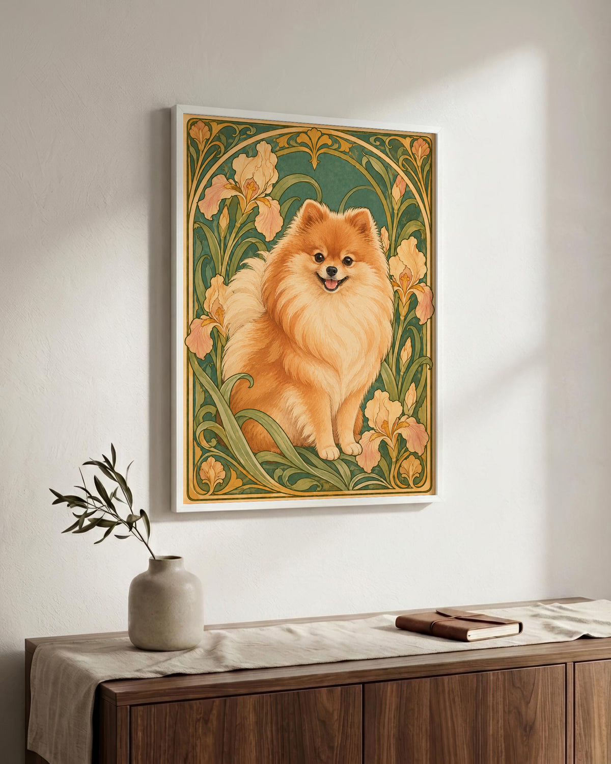 A framed vertical art print featuring a fluffy golden Pomeranian dog in an Art Nouveau floral design, hanging on a light gray wall above a wooden sideboard with a vase and book.
